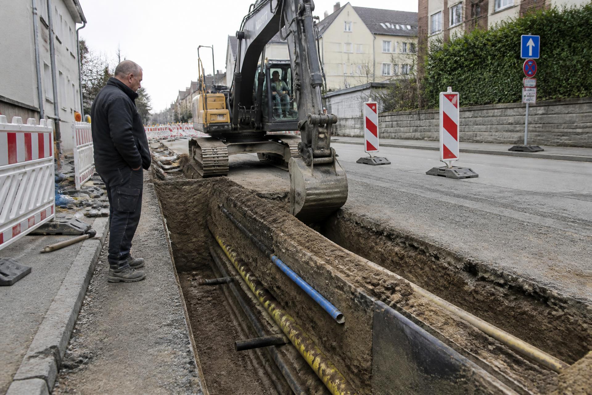 Offener Leitungsgraben in einer Straße: Bagger arbeitet neben freigelegten Strom-, Gas- und Wasserleitungen
